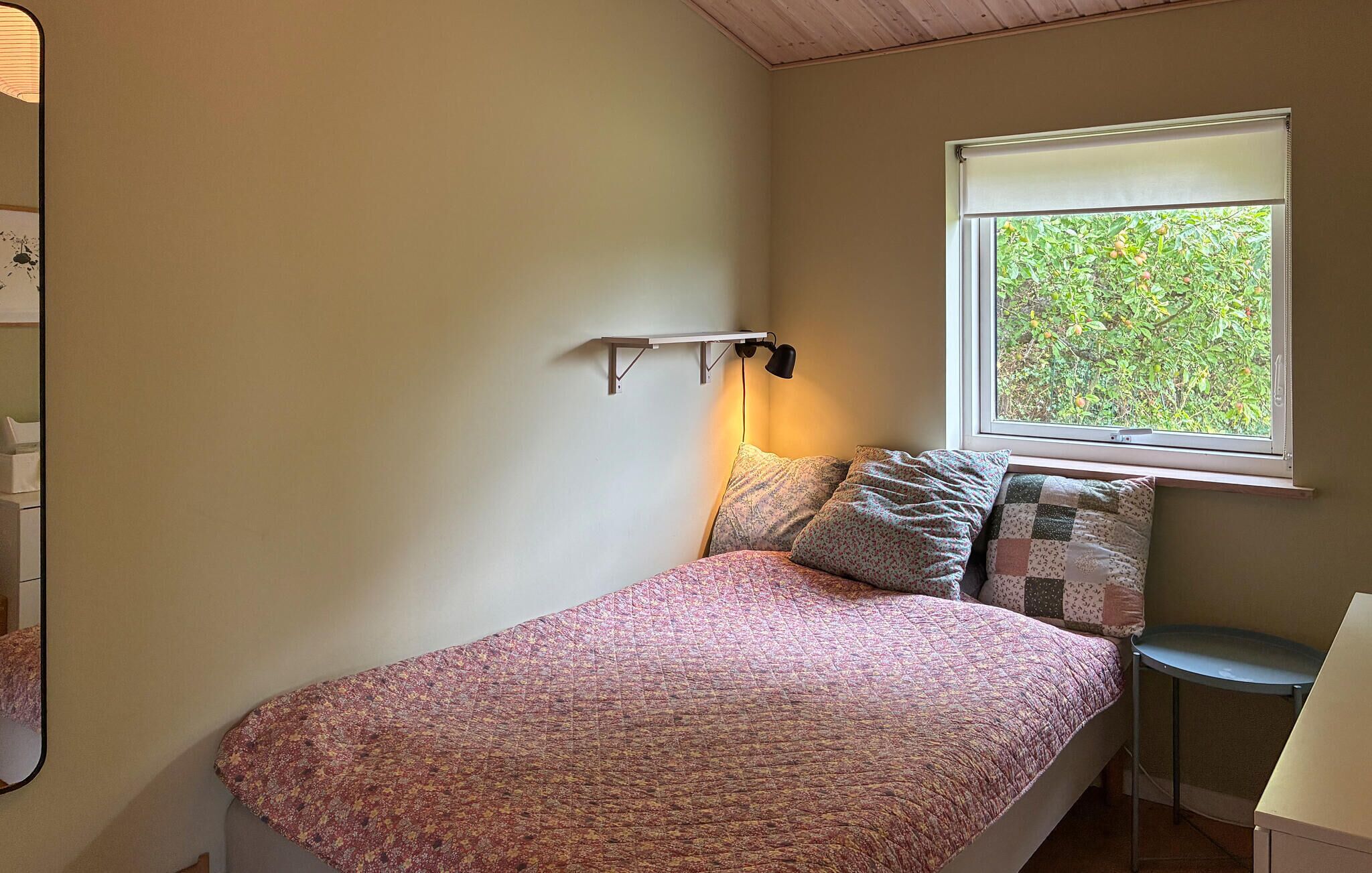 Une chambre à coucher avec un lit, des oreillers, une lampe de chevet, une fenêtre donnant sur un paysage verdoyant et une petite table.