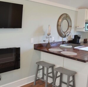 A kitchen with a flat-screen TV mounted on the wall, a round mirror, a coffee maker, and two stools.