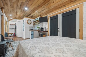 A bedroom with a large bed, wooden ceiling, a desk, and a kitchen area with a clock and shelves.