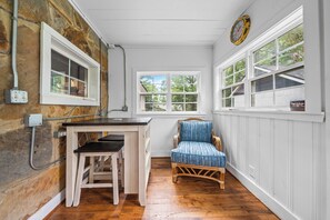 A kitchen with a wooden table and stools, a wicker chair, and a window with a view of trees.