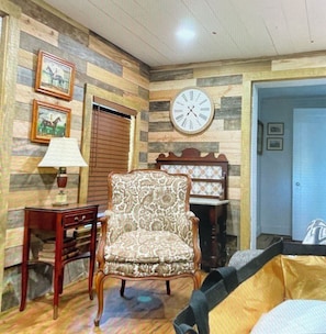 A room with a patterned armchair, a wooden side table with a lamp, a wall clock, and framed pictures.