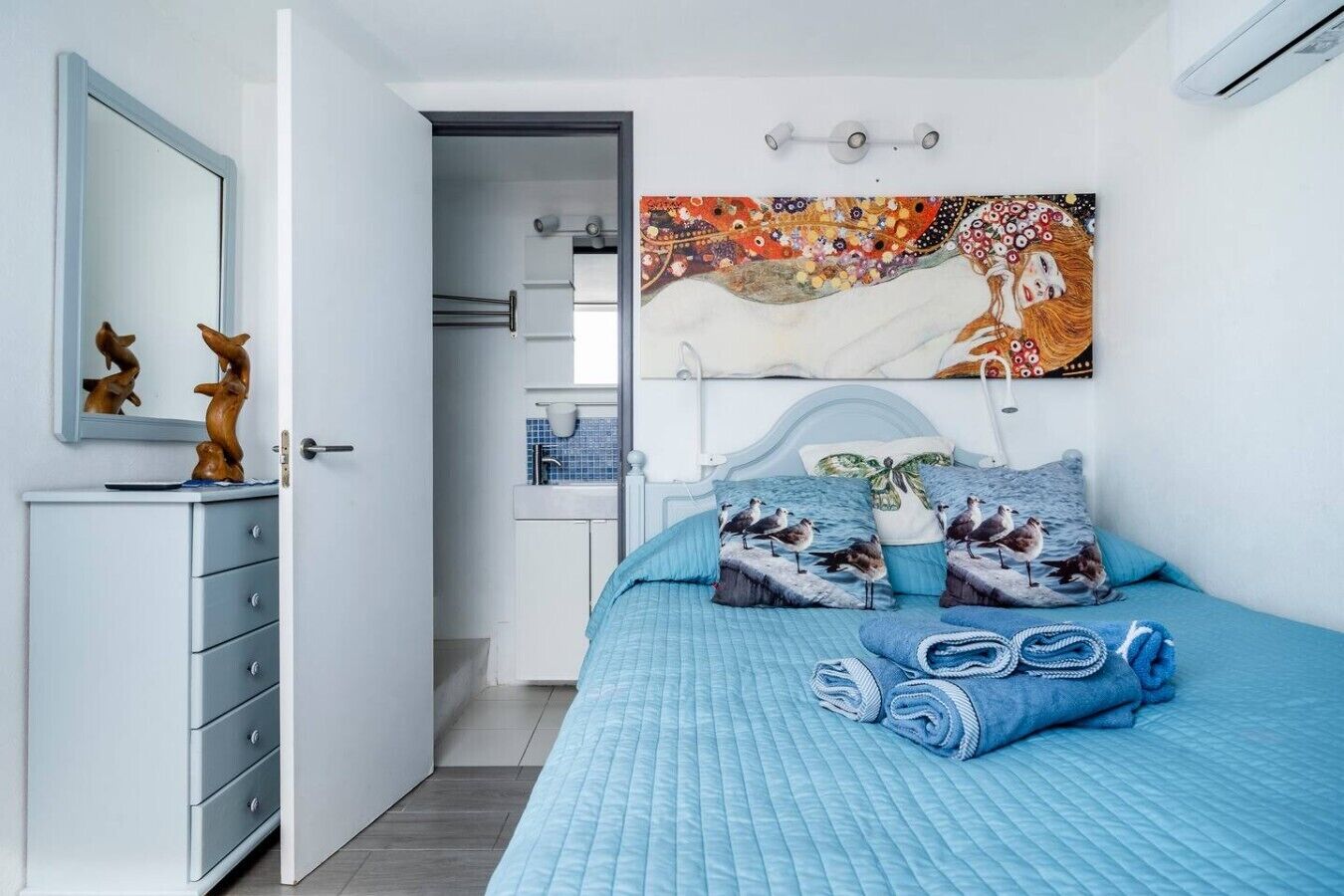 A bedroom with a bed, pillows, a dresser, a mirror, and a wall-mounted artwork.
