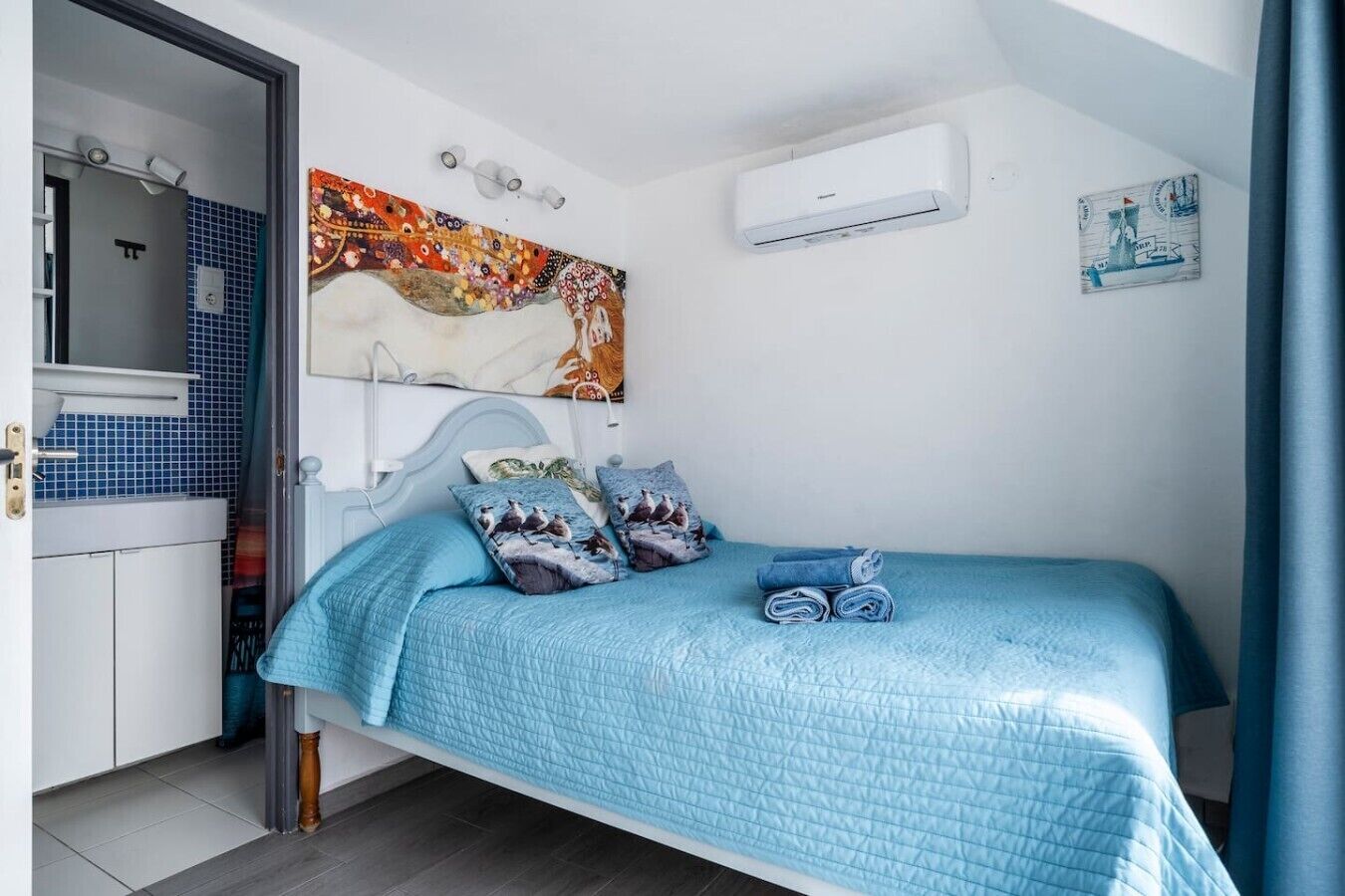Une chambre à coucher avec un lit, des oreillers, un tableau, un climatiseur et une salle de bain visible à travers une porte ouverte.