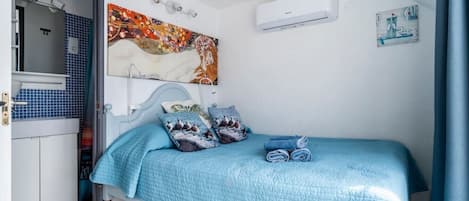 Un dormitorio con cama, almohadas, un cuadro, aire acondicionado y un baño visible a través de una puerta abierta.