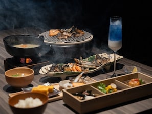 Uma mesa de jantar com diversos pratos, um caldeirão quente e um copo com líquido azul.