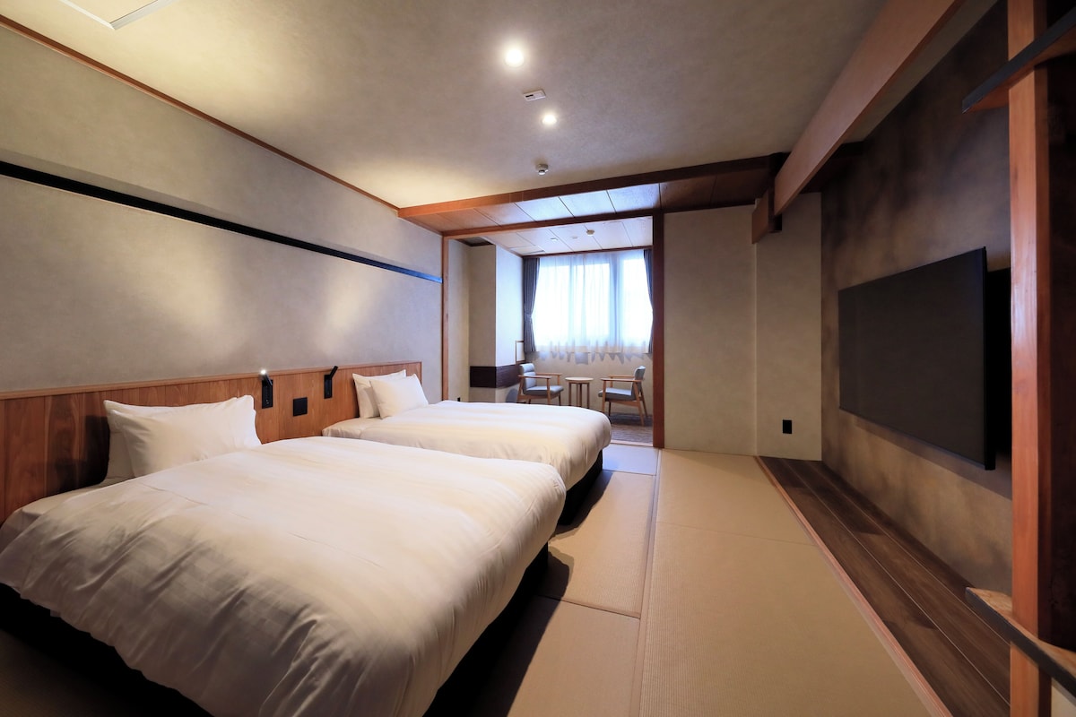 Quarto de hotel com duas camas, área de estar com cadeiras e uma mesa, e uma televisão na parede.