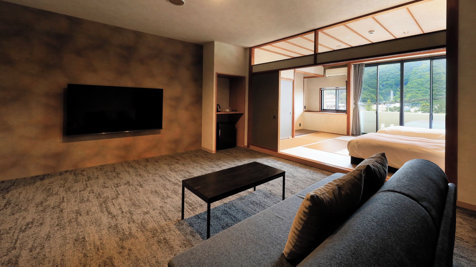 A modern living room with a flat-screen TV, a sofa, a coffee table, and a sliding glass door leading to a balcony with a view of greenery.