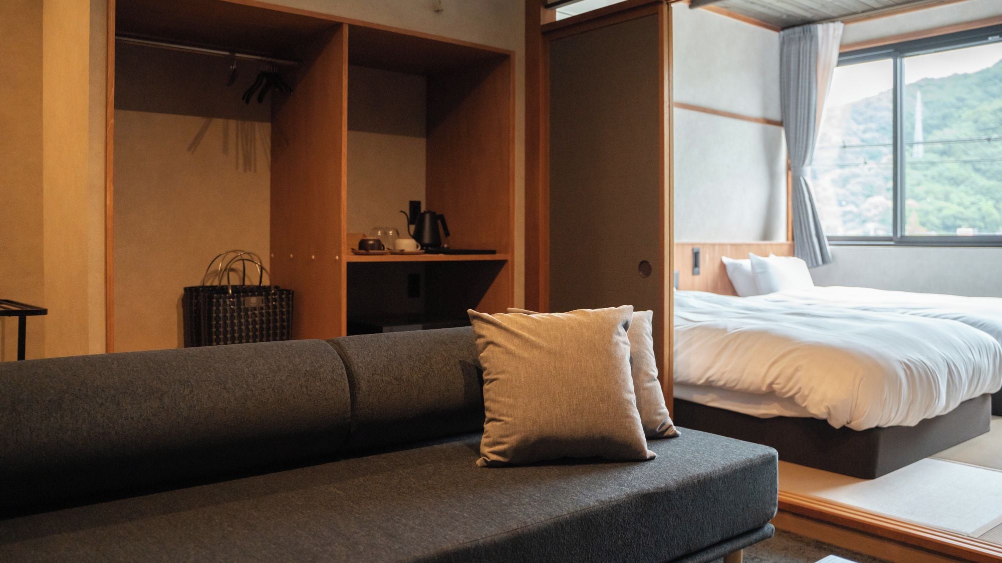 Een moderne kamer met een grijze bank, een bed met witte beddengoed, een houten garderobe en uitzicht op groen door een raam.
