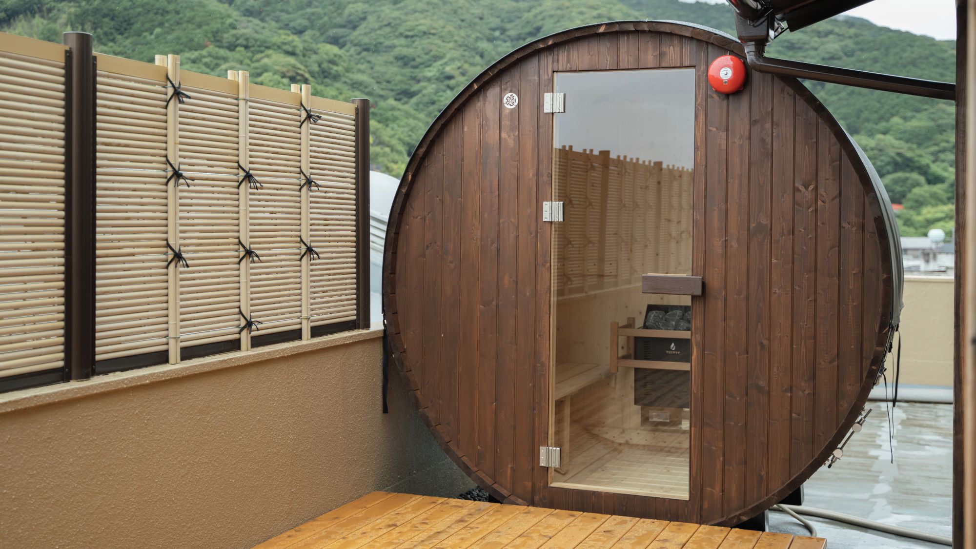 A wooden sauna with a sliding door, a small wooden bench inside, and a view of a forested hillside.