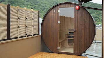 A wooden sauna with a sliding door, a small wooden bench inside, and a view of a forested hillside.