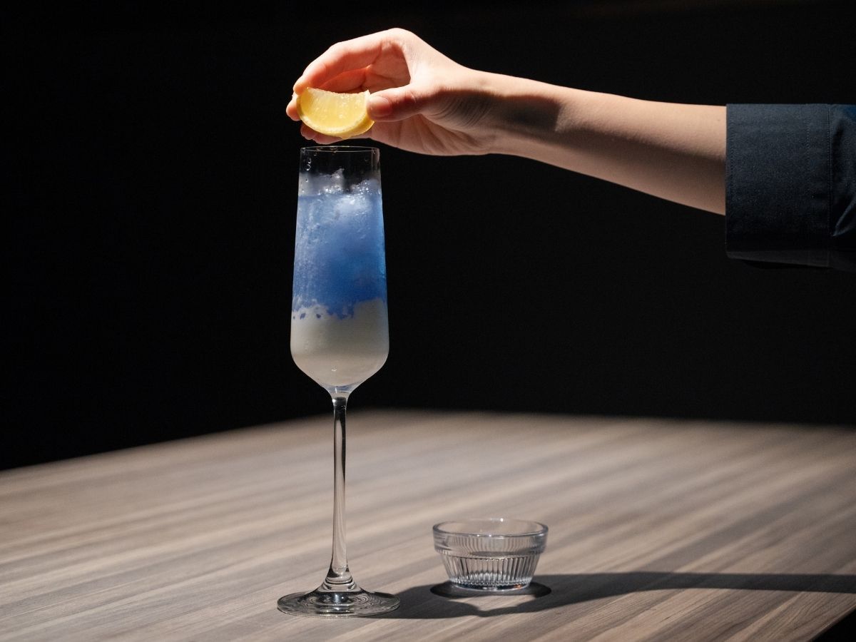 Une main tenant une tranche de citron au-dessus d’un grand verre contenant un liquide bleu, le tout posé sur une surface en bois, avec un petit verre à côté.