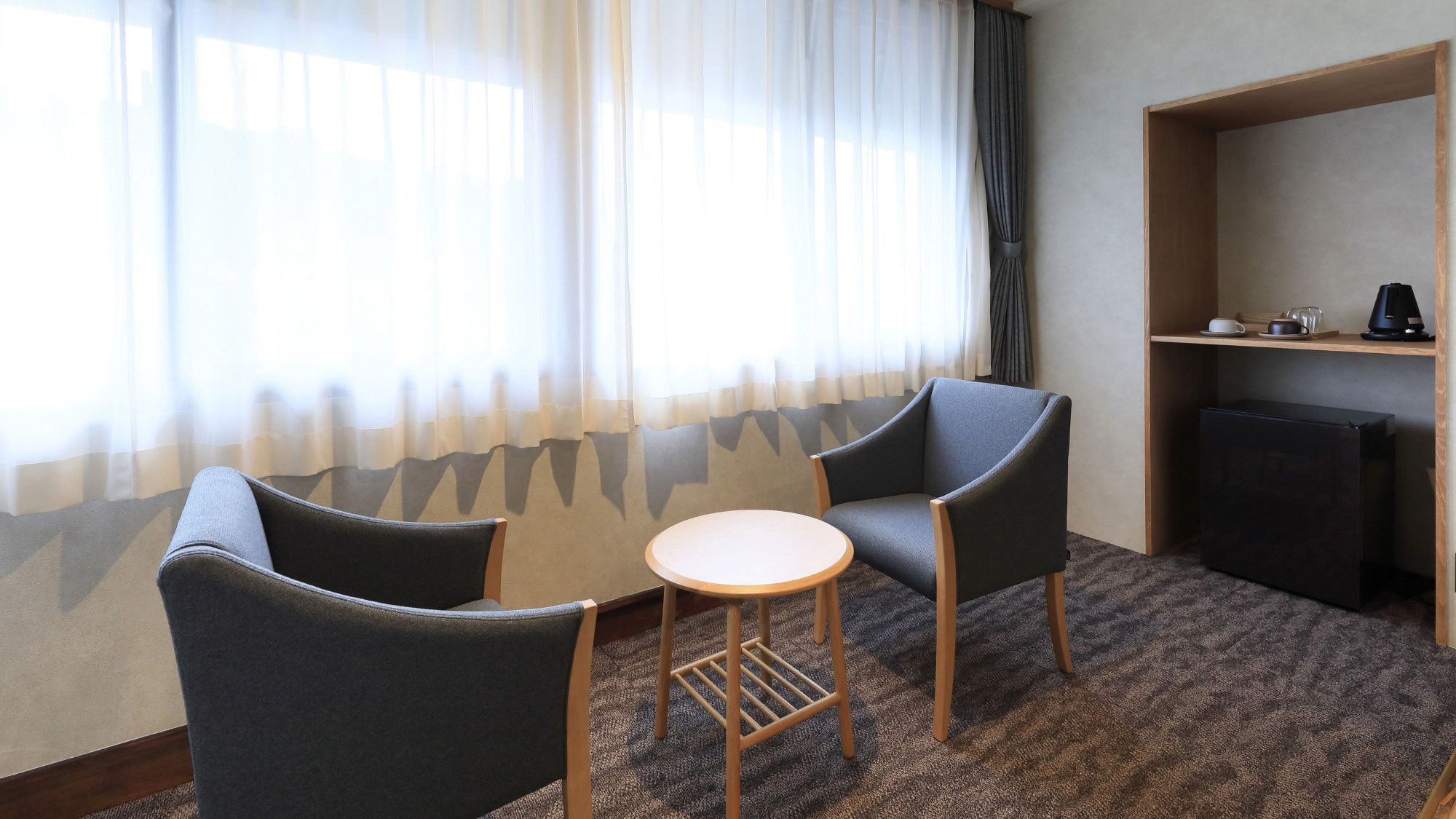 Ein Hotelzimmer mit zwei Sesseln, einem kleinen runden Tisch, einem Bücherregal und einem Fenster mit durchsichtigen Vorhängen.
