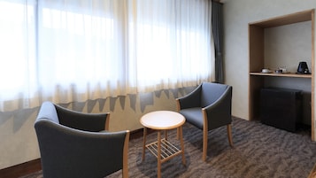 Habitación de hotel con dos sillones, una mesita redonda, una estantería y un ventanal con cortinas translúcidas.