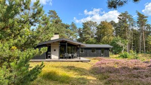 Uma cabana moderna com varanda coberta e um edifício separado, rodeada por uma floresta densa e urzes.