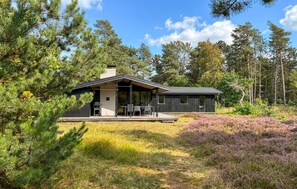 Eine moderne Holzhütte mit überdachter Terrasse und einem separaten Gebäude, umgeben von dichtem Wald und Heidekraut.