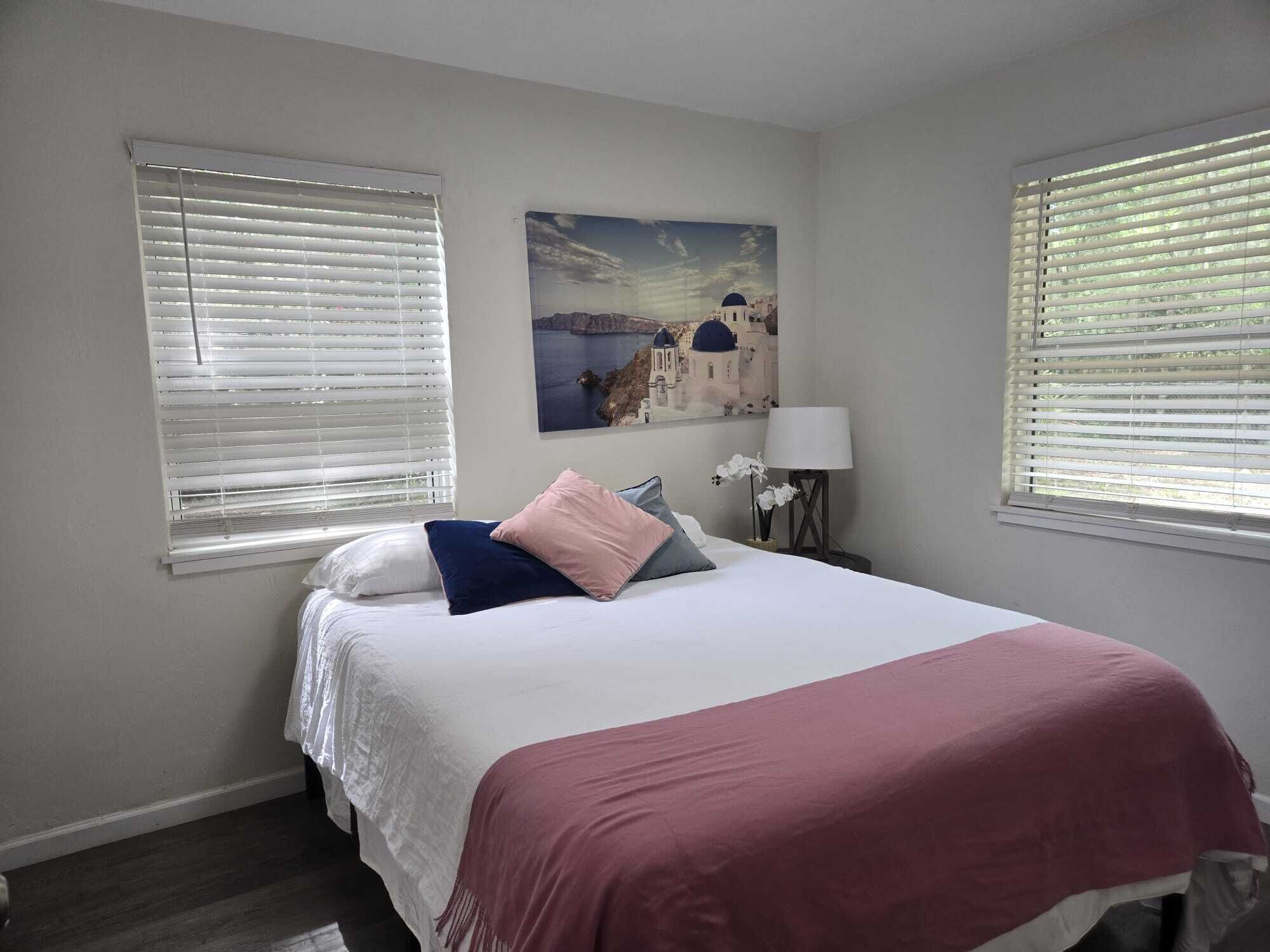A bedroom with a bed, two windows with blinds, a nightstand with a lamp, and a painting of a coastal scene.