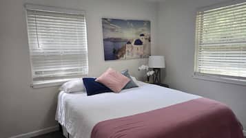 A bedroom with a bed, two windows with blinds, a nightstand with a lamp, and a painting of a coastal scene.