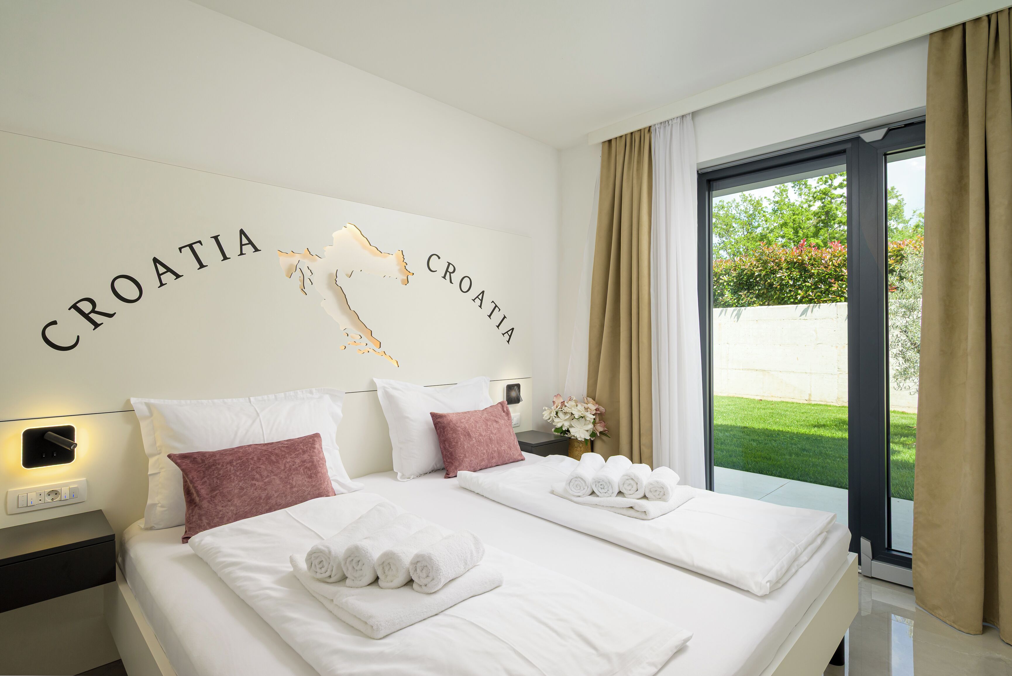 A hotel room with a bed, pillows, a nightstand, a wall decoration, and a view of greenery outside.