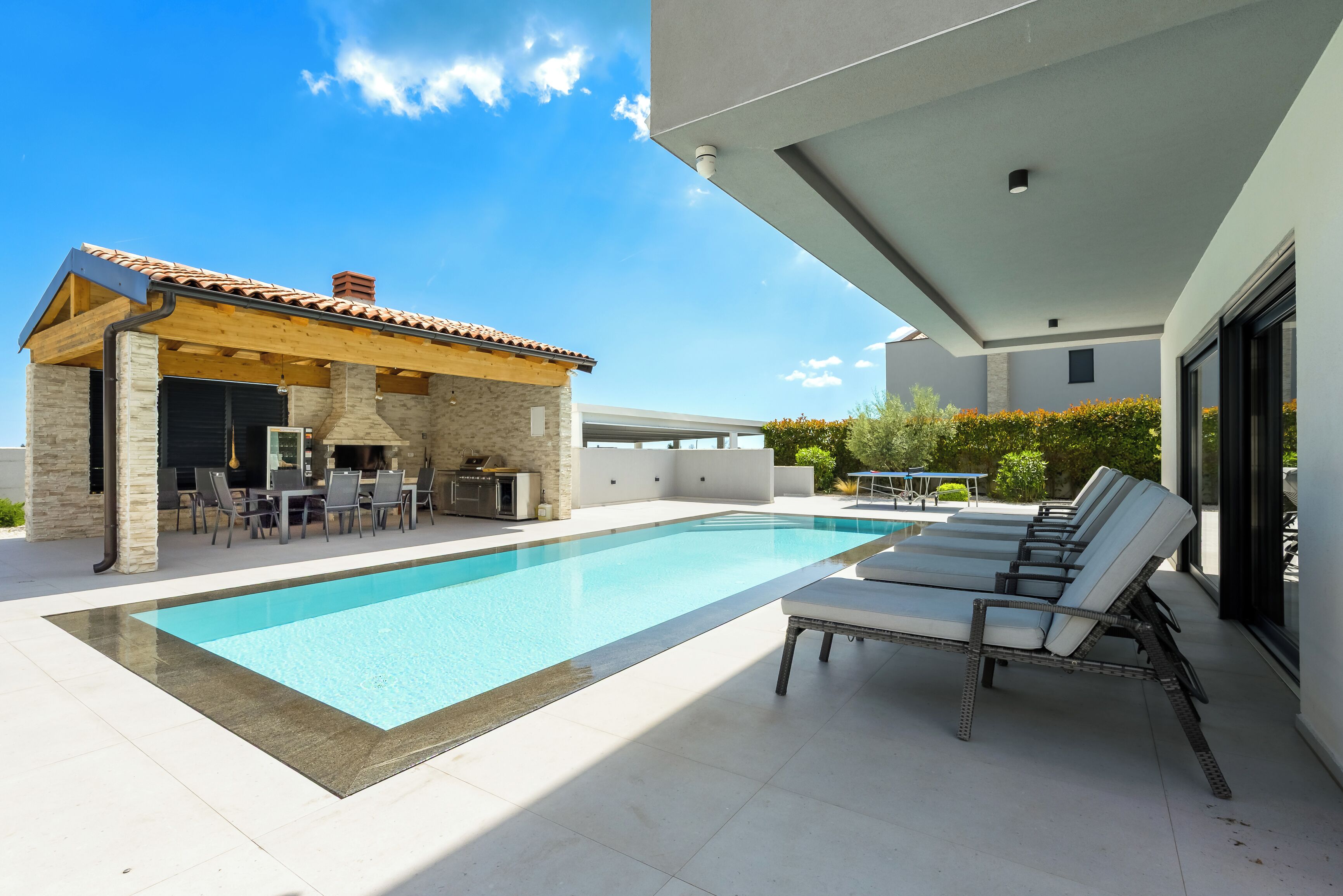 Un'area piscina moderna all'aperto con una veranda coperta, lettini e un angolo barbecue.