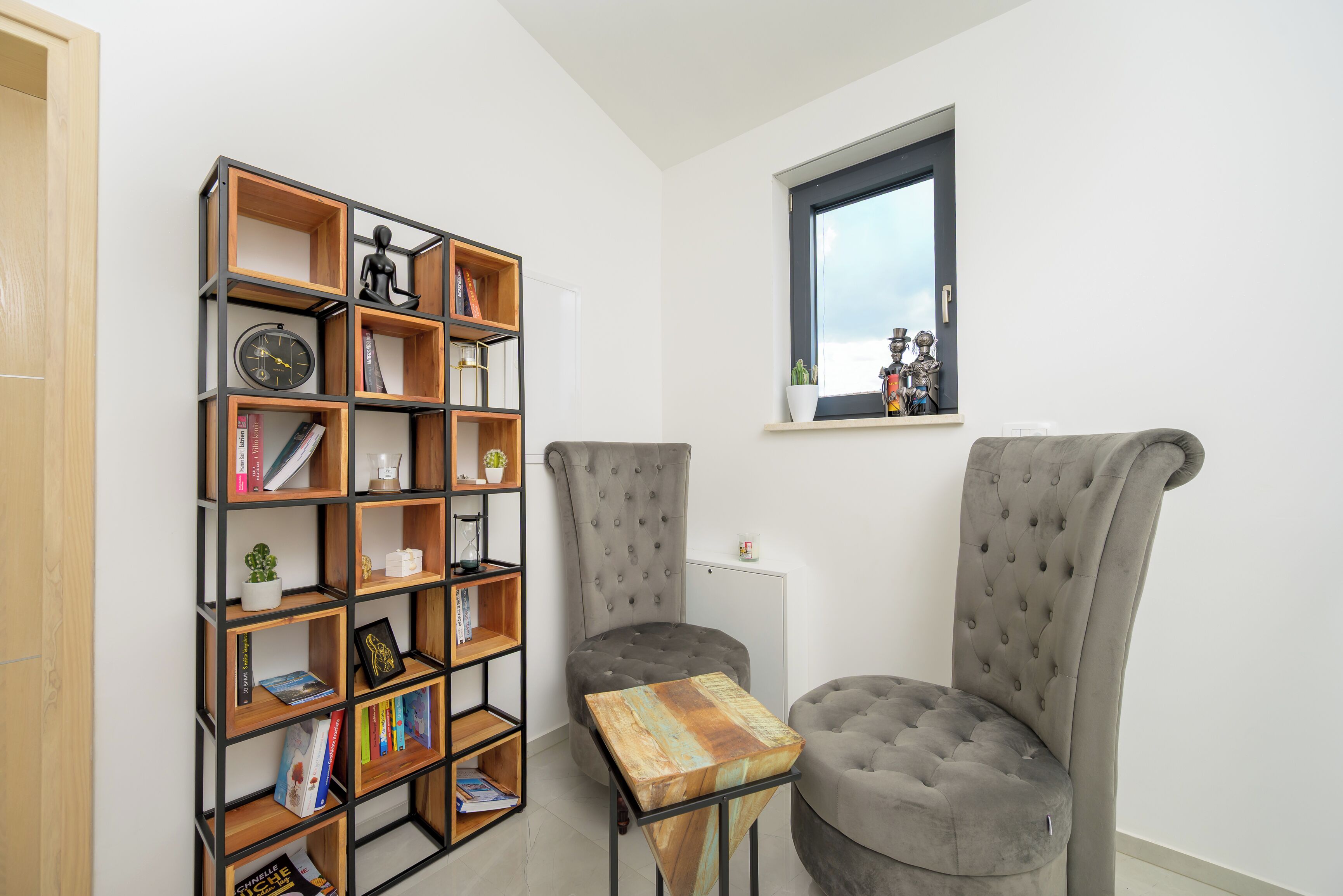 A room with a bookshelf, two grey upholstered chairs, a small table, and a window with a view.