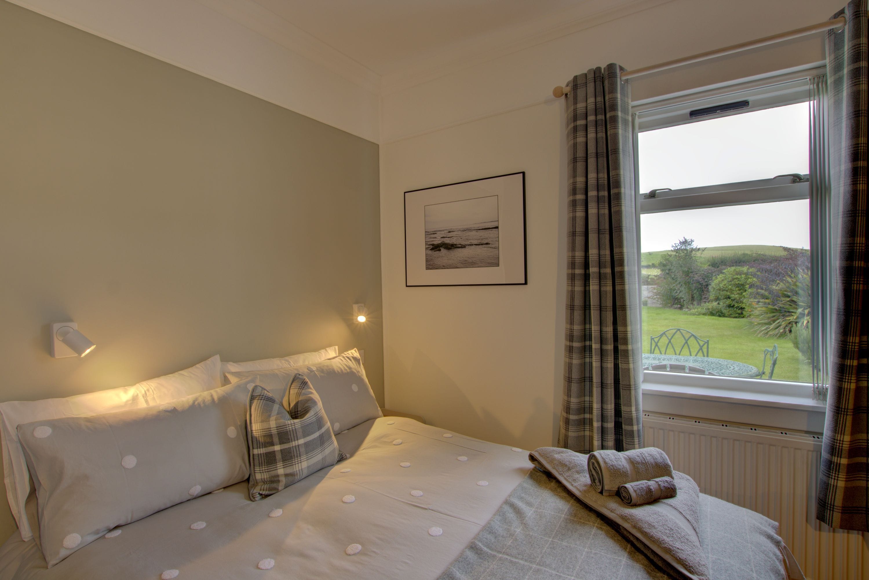 A bedroom with a bed, pillows, a framed picture, a window with curtains, and a view of the outdoors.