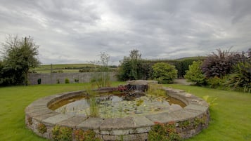 A circular stone pond with a small waterfall, surrounded by grass and various plants.
