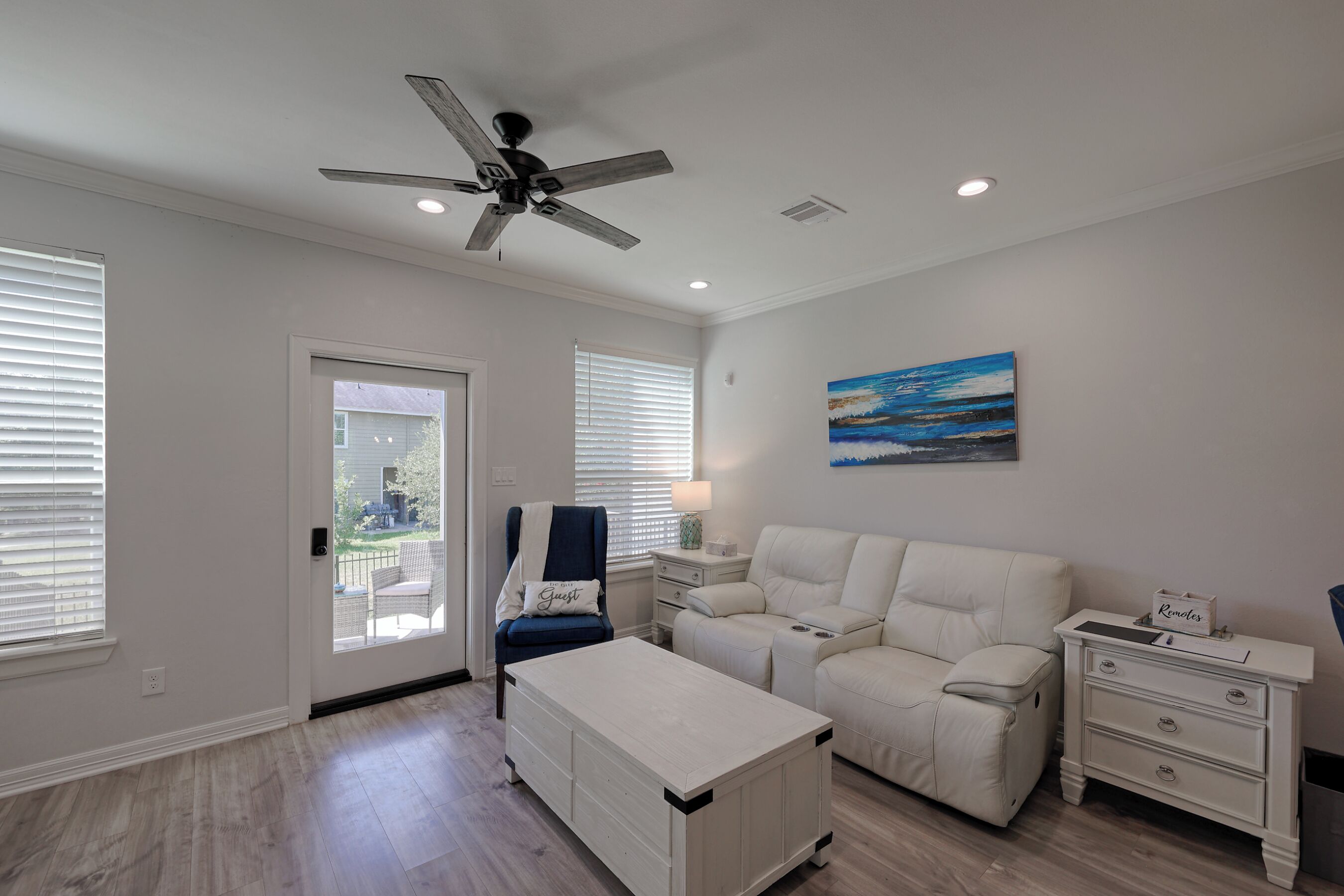 A spacious living room with a ceiling fan, a painting, a white sofa, a recliner, a coffee table, and a side table with a lamp.
