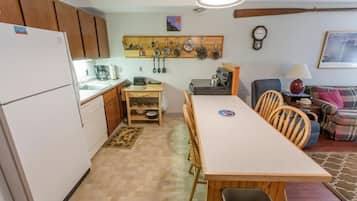 A compact kitchen with wooden cabinets, a dining table, and chairs. A coffee maker and kettle are on the counter.