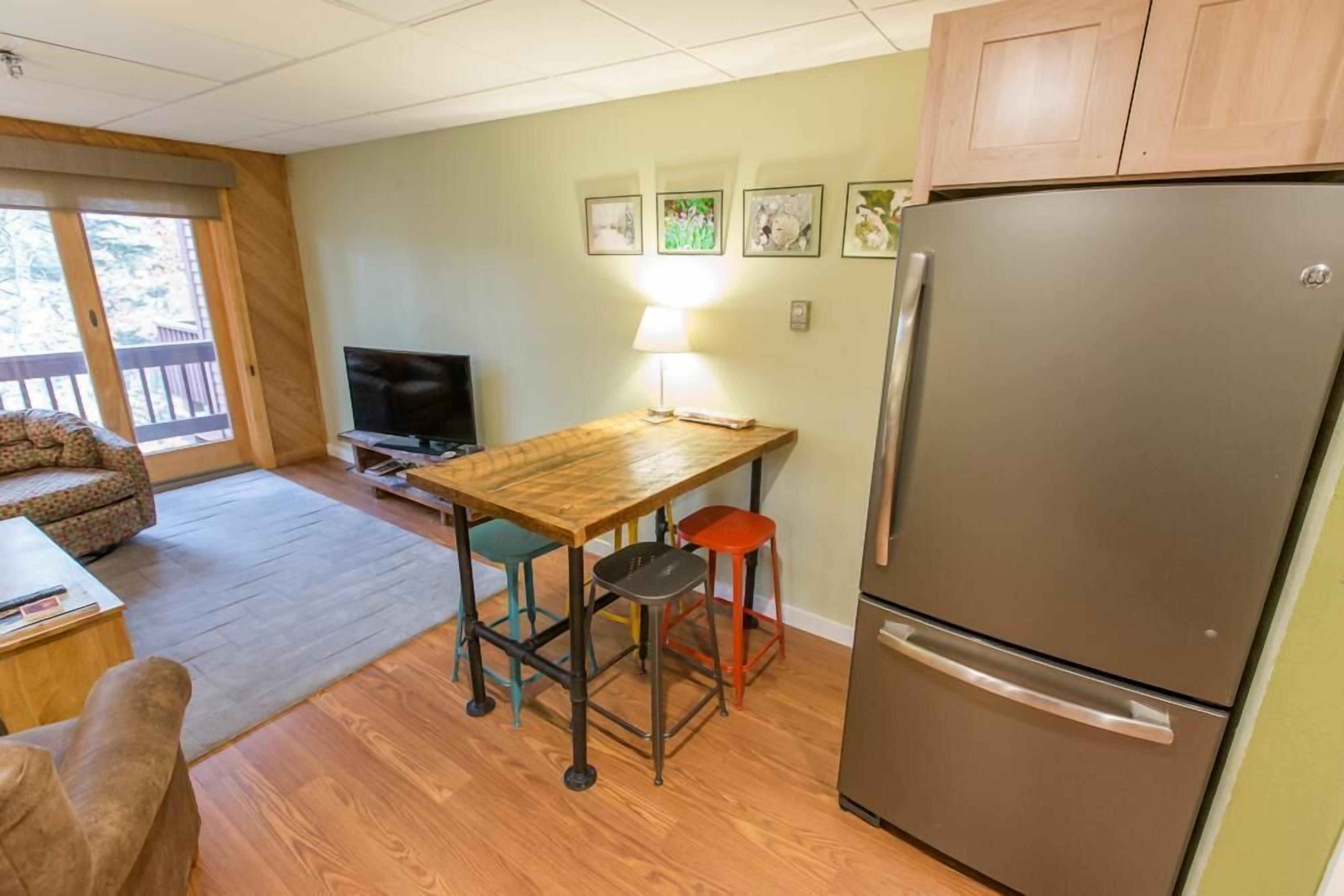 A compact kitchen with a refrigerator, wooden table, and colorful stools.