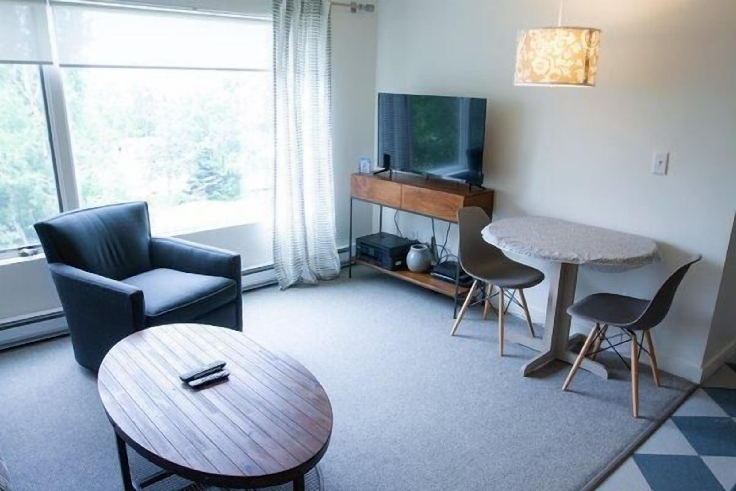 A modern hotel room with a sofa, a round table, a TV, and a dining area.