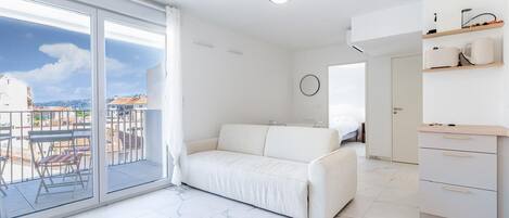 Uma sala de estar moderna com um sofá branco, uma varanda com vista para edifícios e uma área de cozinha com armários brancos e bancada.