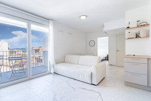 Ein modernes Wohnzimmer mit einem weißen Sofa, einem Balkon mit Blick auf Gebäude und einer Küchenzeile mit weißen Schränken und einer Arbeitsfläche.