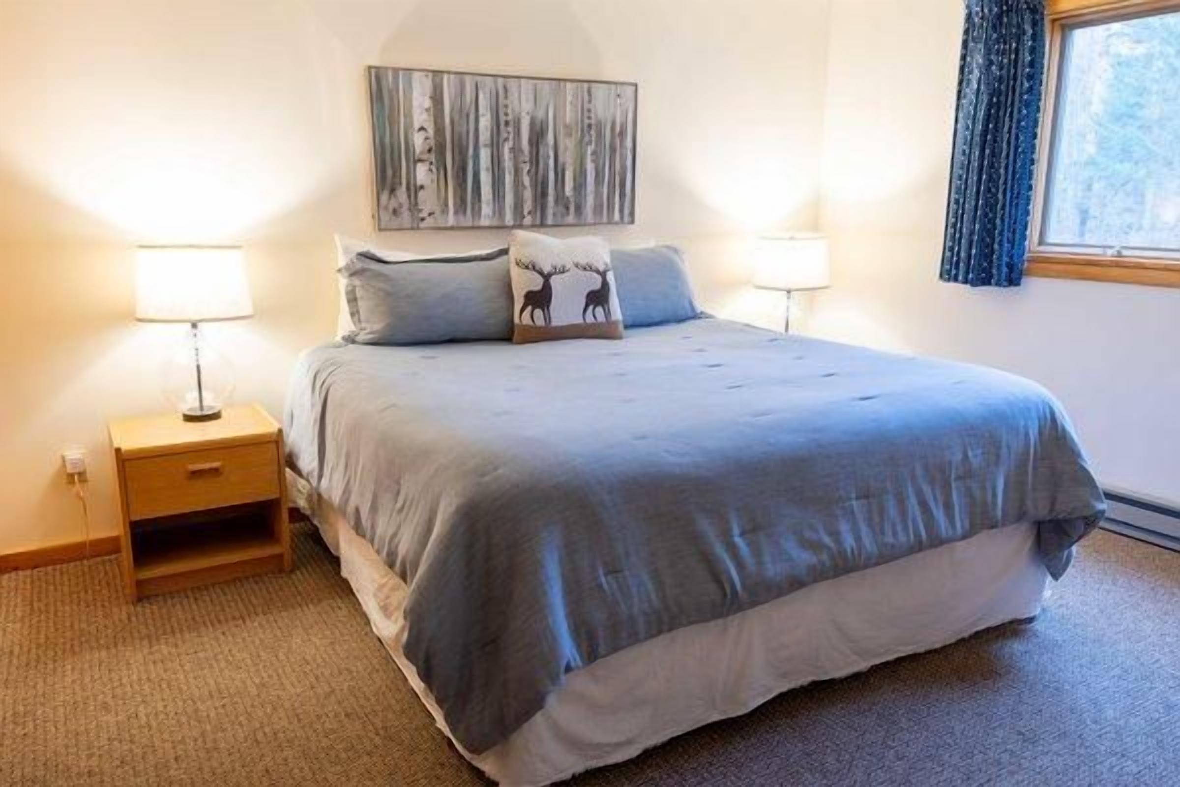 A bedroom with a bed, two bedside tables, a nightstand, a wall-mounted artwork, and a window with curtains.