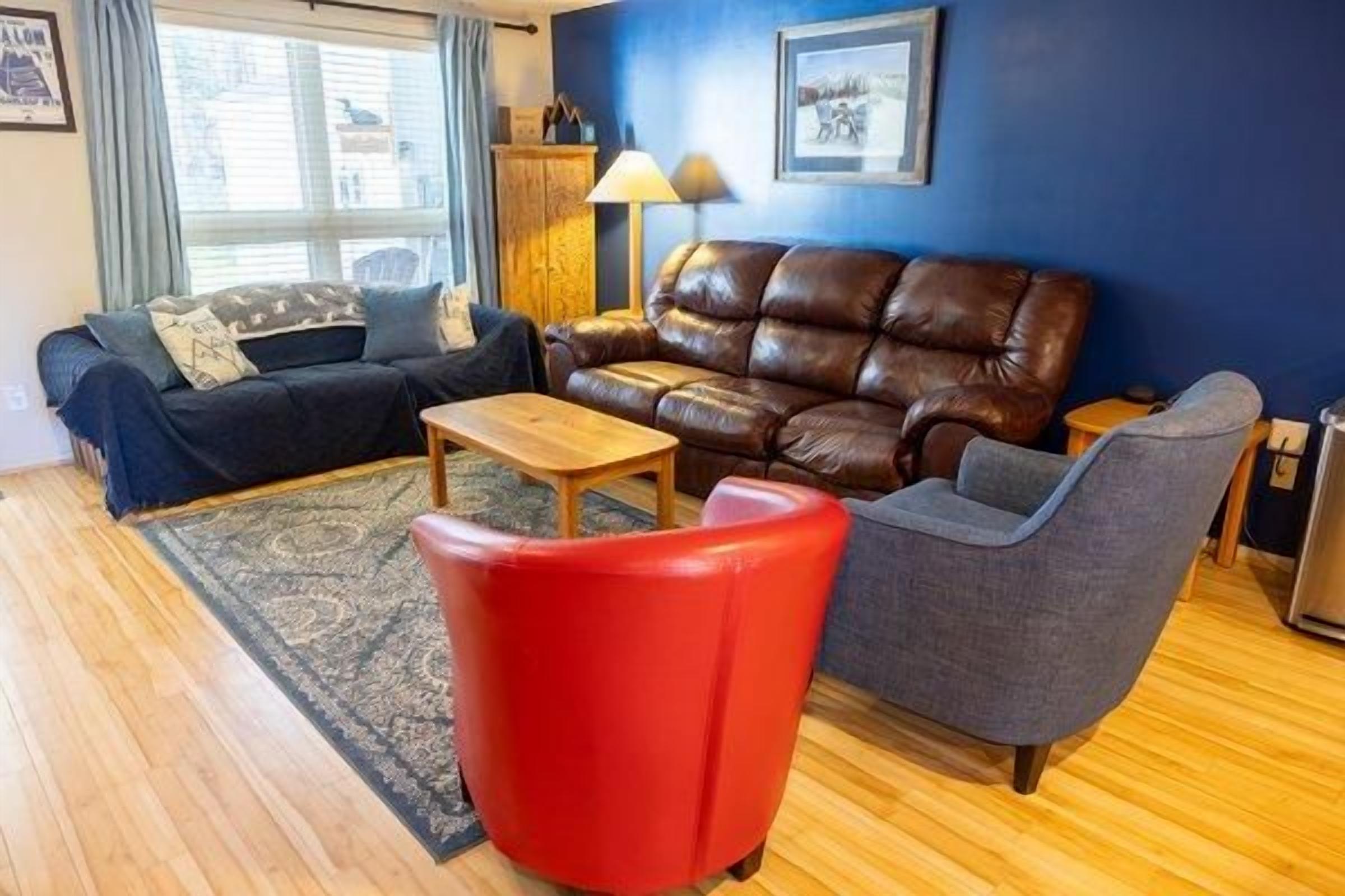 Ein Wohnzimmer mit einem braunen Ledersofa, einem roten Sessel, einem grauen Ohrensessel, einem Holztisch und einer blauen Wand.