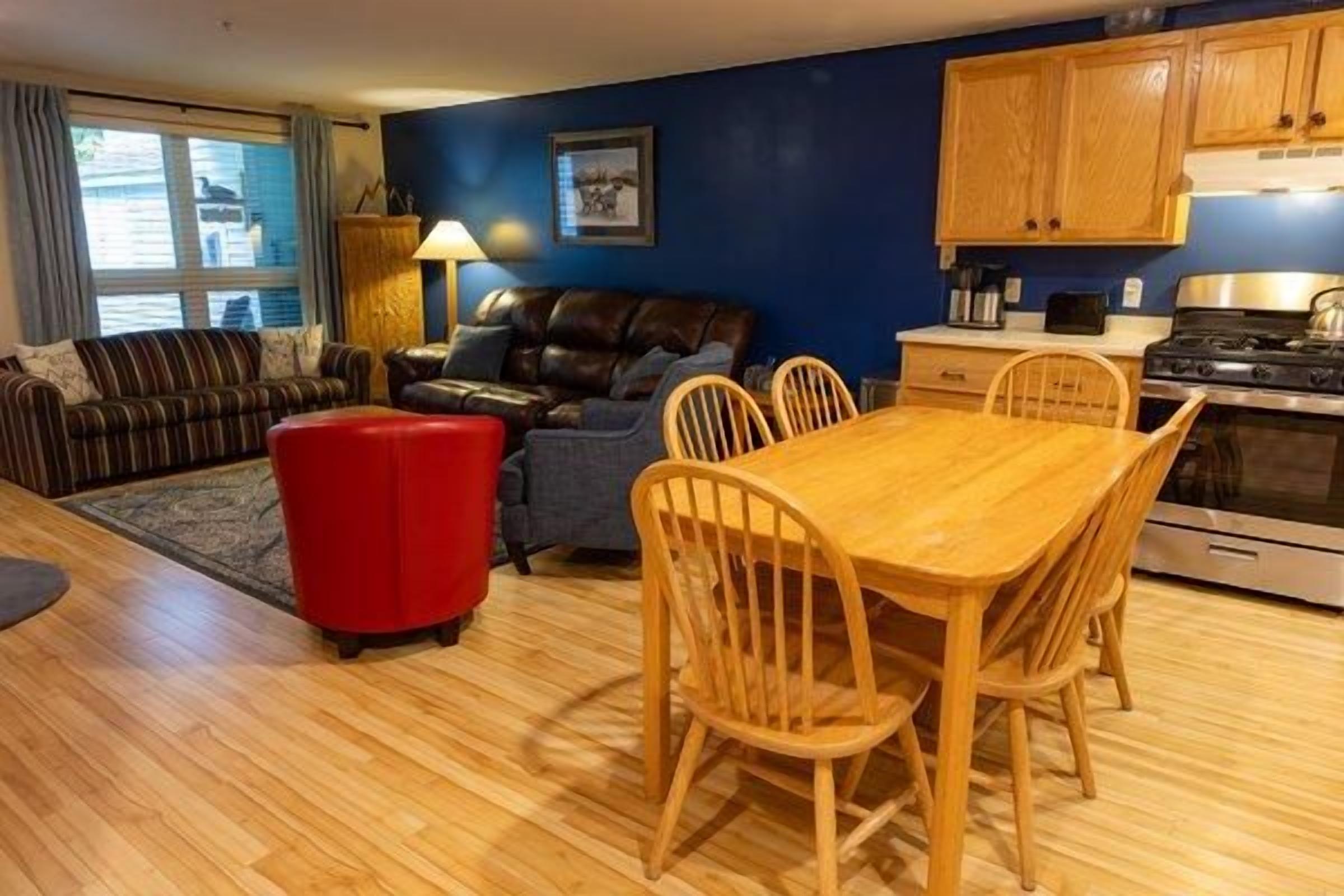 Un salon comprenant un canapé, un pouf rouge, une table à manger avec des chaises, et un coin cuisine avec des placards et des appareils électroménagers.