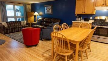 Un salon comprenant un canapé, un pouf rouge, une table à manger avec des chaises, et un coin cuisine avec des placards et des appareils électroménagers.
