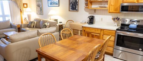 A cozy living room with a sofa, a wooden dining table, and chairs. There is a kitchen area with a stove, microwave, and cabinets.