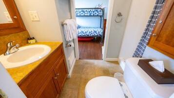 A compact bathroom with a sink, toilet, and a small mirror. There is a visible towel rack and a shower area in the background.
