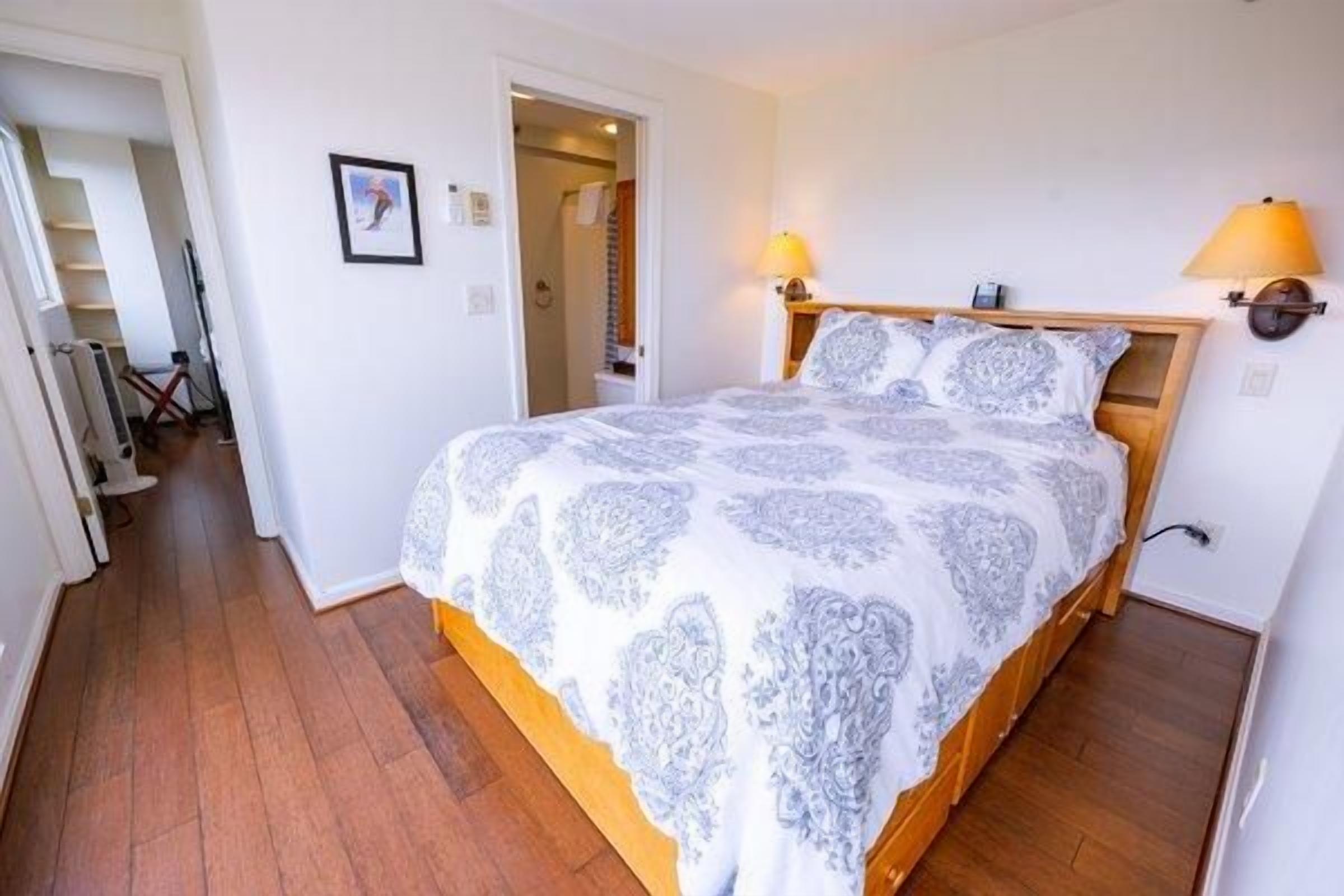 A bedroom with a bed, wooden headboard, bedside lamps, and a door leading to a bathroom.