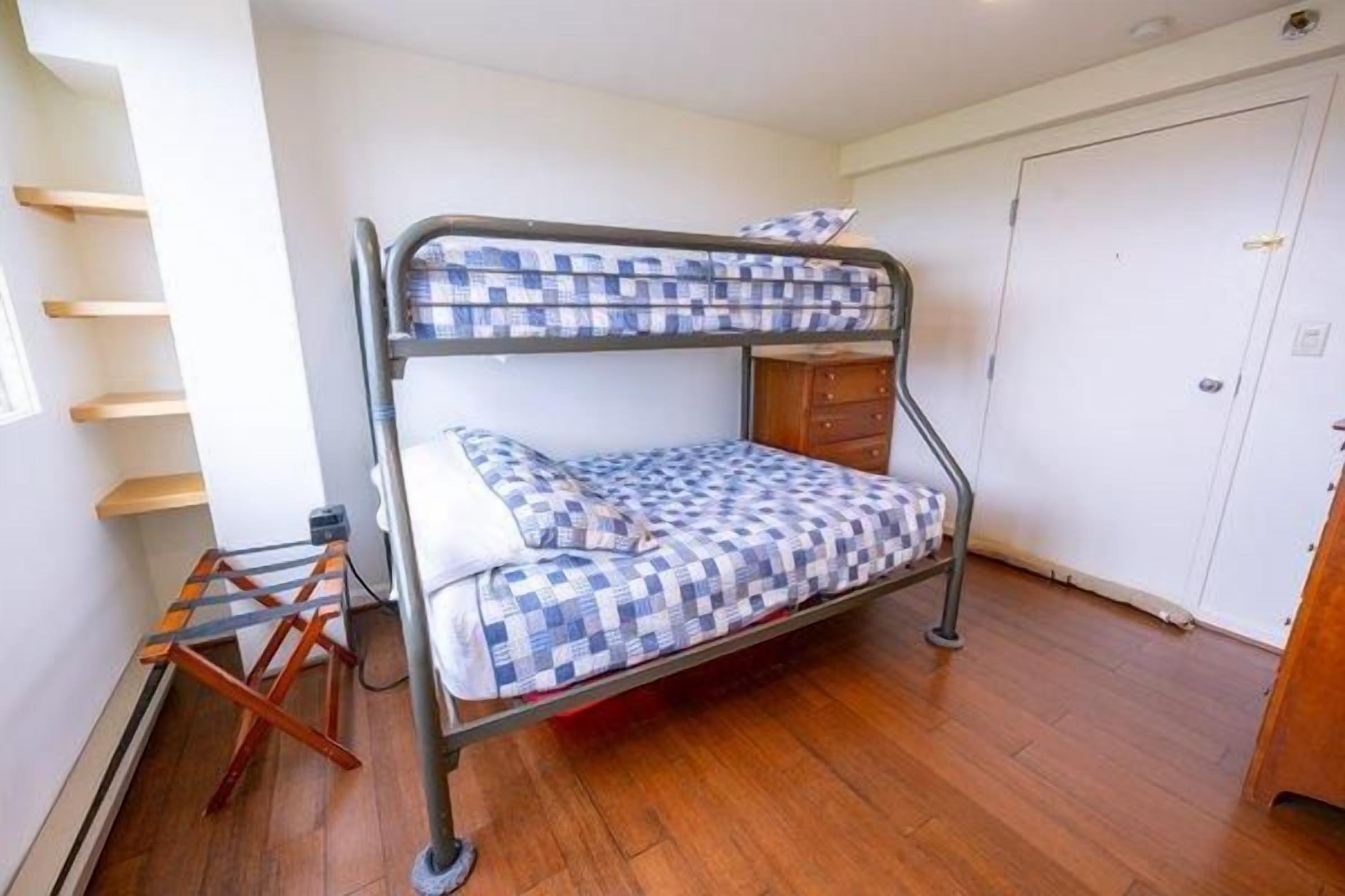 Ein Stockbett mit kariertem Bettbezug, eine Holzkommode und eine weiße Tür in einem Zimmer mit Holzboden.