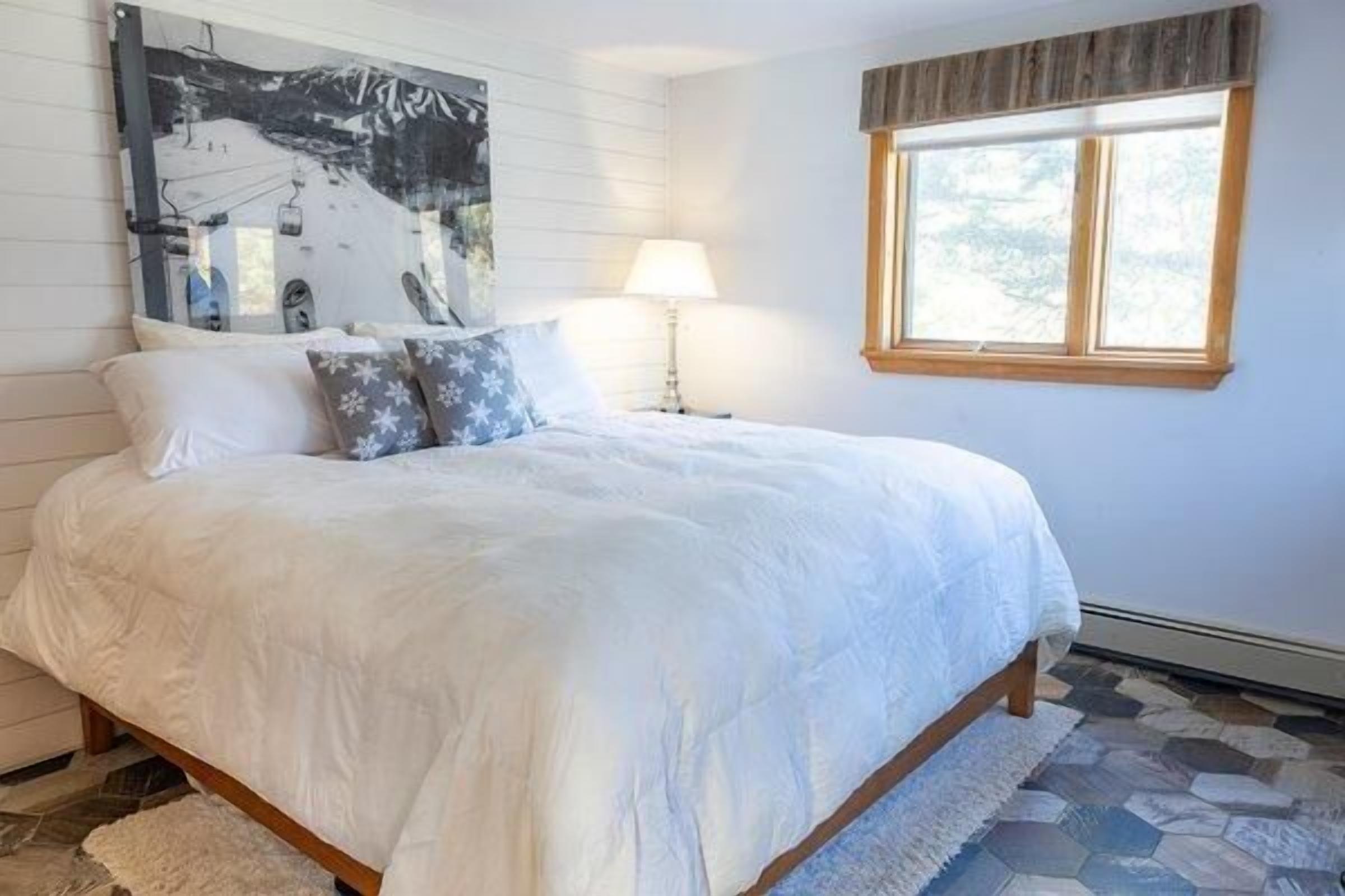 A bedroom with a large bed, a nightstand, a lamp, a window with blinds, and a painting on the wall.