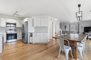 A modern kitchen with stainless steel appliances, white cabinetry, and a wooden dining table with white chairs. - Wavecrest Cottage-Sleeps 14 Beach (Laketown Township)