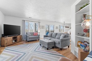 A modern living room with a flat-screen TV, grey sofas, a patterned rug, and a bookshelf with various items. - Wavecrest Cottage-Sleeps 14 Beach (Laketown Township)