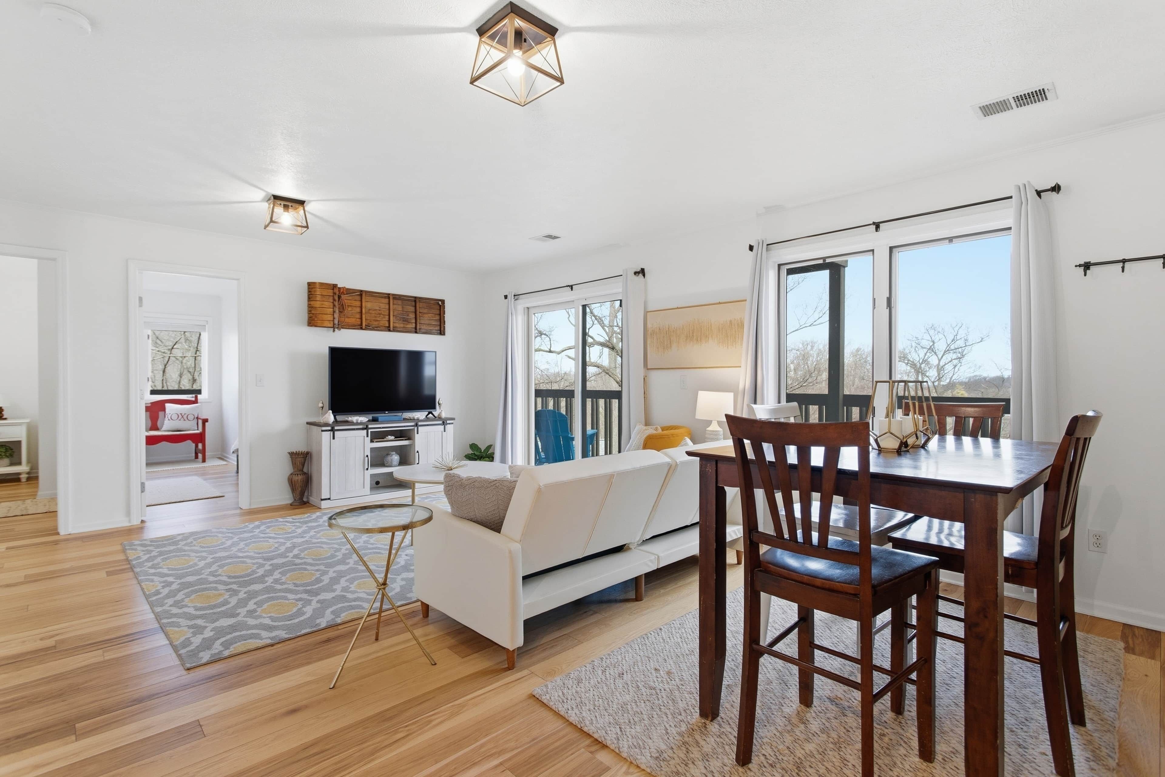 A modern living room with a flat-screen TV, a dining table with chairs, a sofa, and a large window.