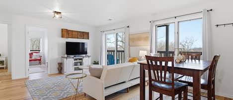 A modern living room with a flat-screen TV, a dining table with chairs, a sofa, and a large window.