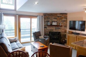 Ein Wohnzimmer mit einer Couch, Sesseln, einem Couchtisch, einem Kamin, einem Fernseher und einer Schiebetür zu einem Außenbereich.