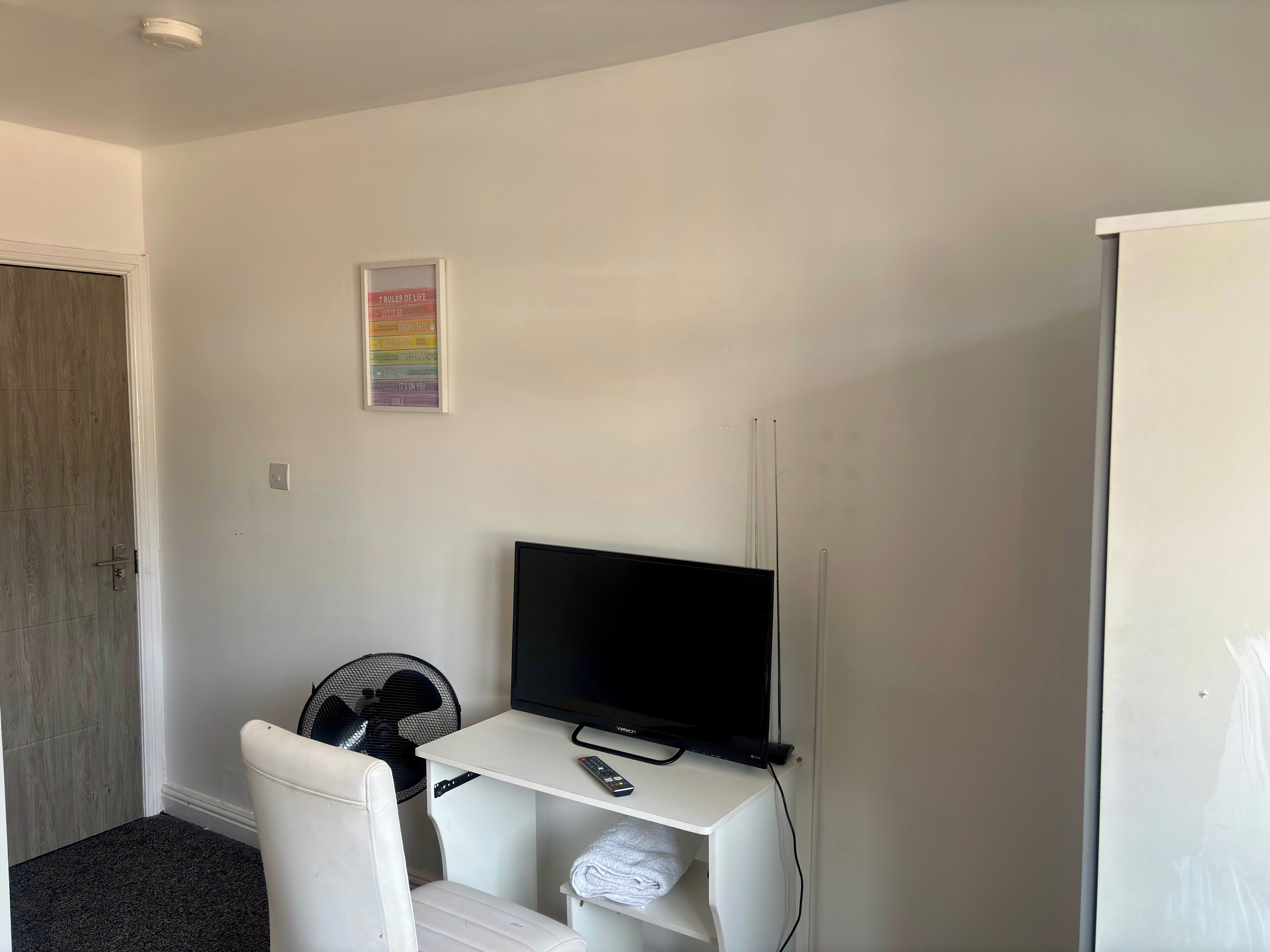 Ein Zimmer mit weißem Schreibtisch, einem Flachbildfernseher, einem Ventilator und einem gerahmten Bild an der Wand.