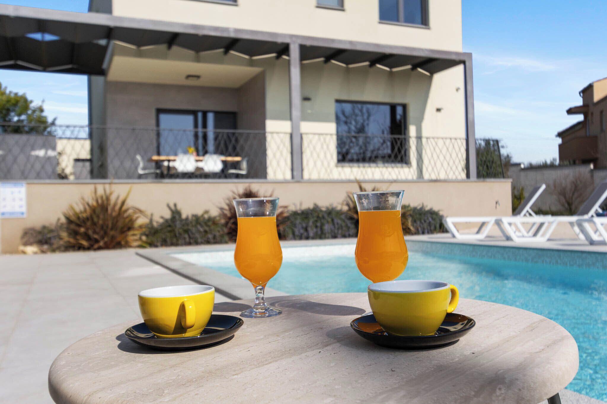 Mesa à beira da piscina com dois copos amarelos e dois copos com bebida de laranja, uma piscina e um prédio com alpendre ao fundo.