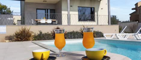 Mesa à beira da piscina com dois copos amarelos e dois copos com bebida de laranja, uma piscina e um prédio com alpendre ao fundo.