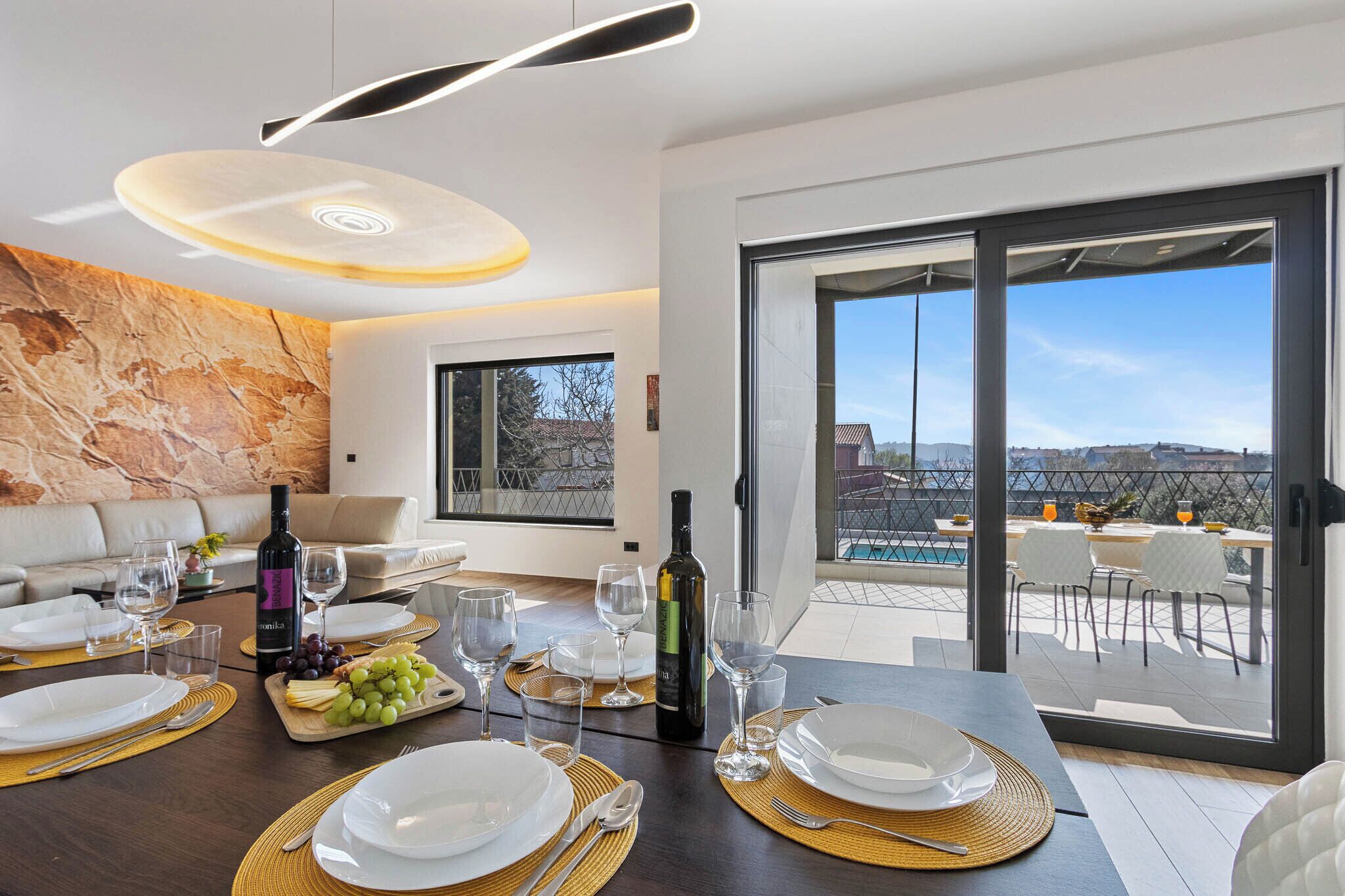 A modern dining area with a table set for four, a wine bottle, and a view of a pool and buildings.